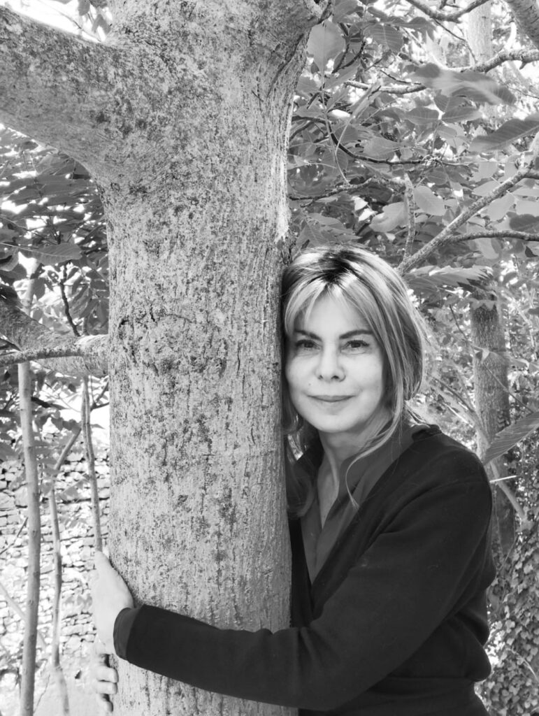 Foto in bianco e nero che ritrae Antonella Anedda in un parco che abbraccia il tronco di un albero