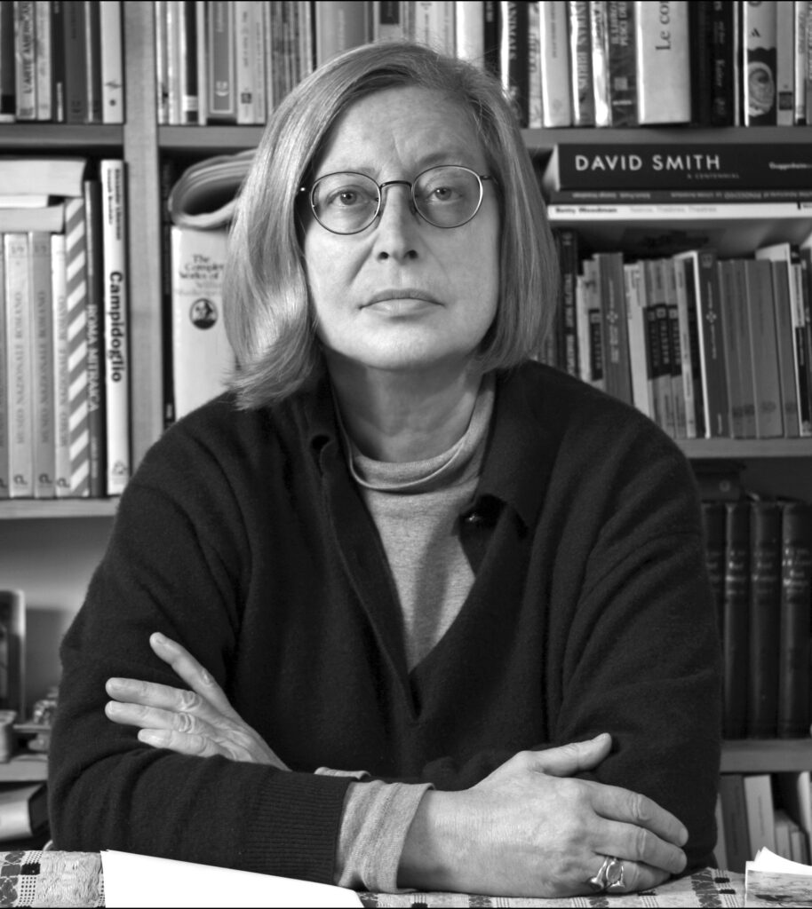 Patrizia Cavalli seduta di fronte a una libreria della sua casa